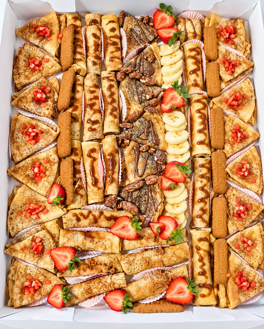 Assorted rolled and folded handheld crêpes garnished with strawberries, bananas, sauces, nuts, and biscuits